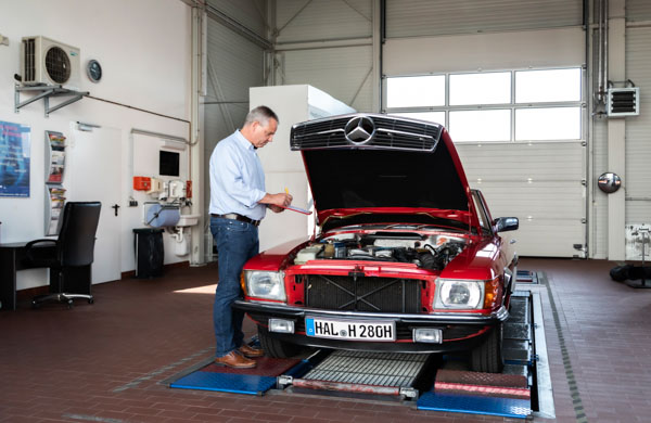 Kfz-Sachverständiger steht neben offener Motorhaube eines roten Autos und notiert auf einem Block